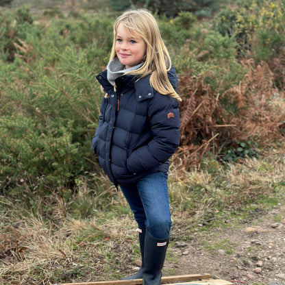A young girl stands on a wooden sled outdoors in a grassy, wooded setting. She is wearing a navy blue puffer jacket, a gray knit scarf, blue jeans, and black rubber boots.