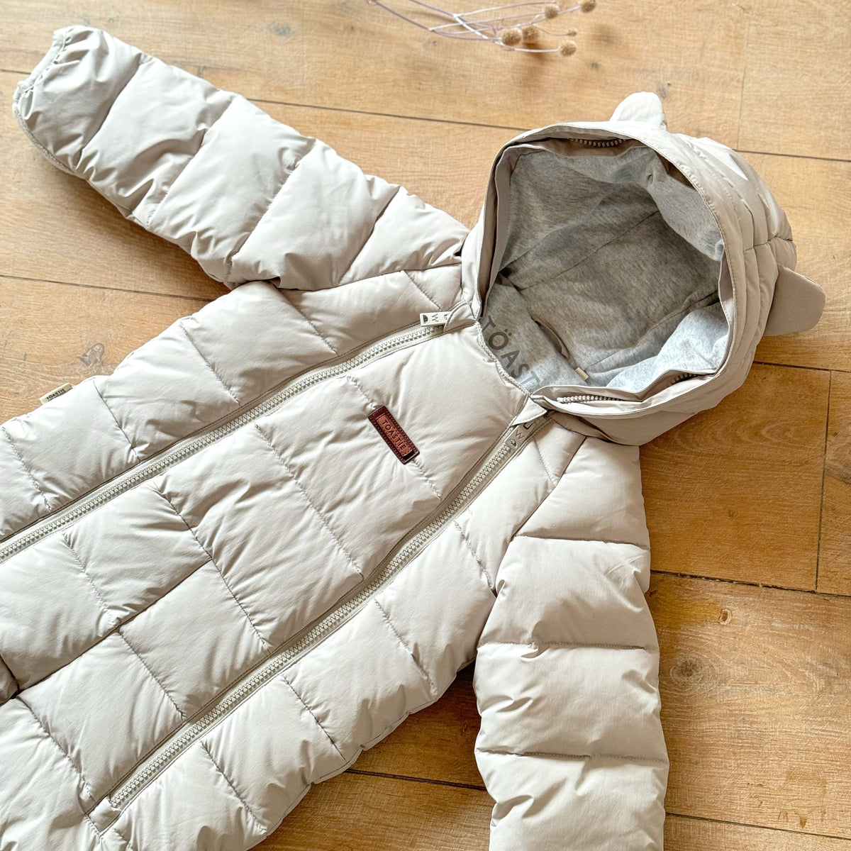 A white hooded snowsuit laid flat on a wooden floor, with the hood open to show a gray inner lining. A small brown brand tag is visible on the chest.