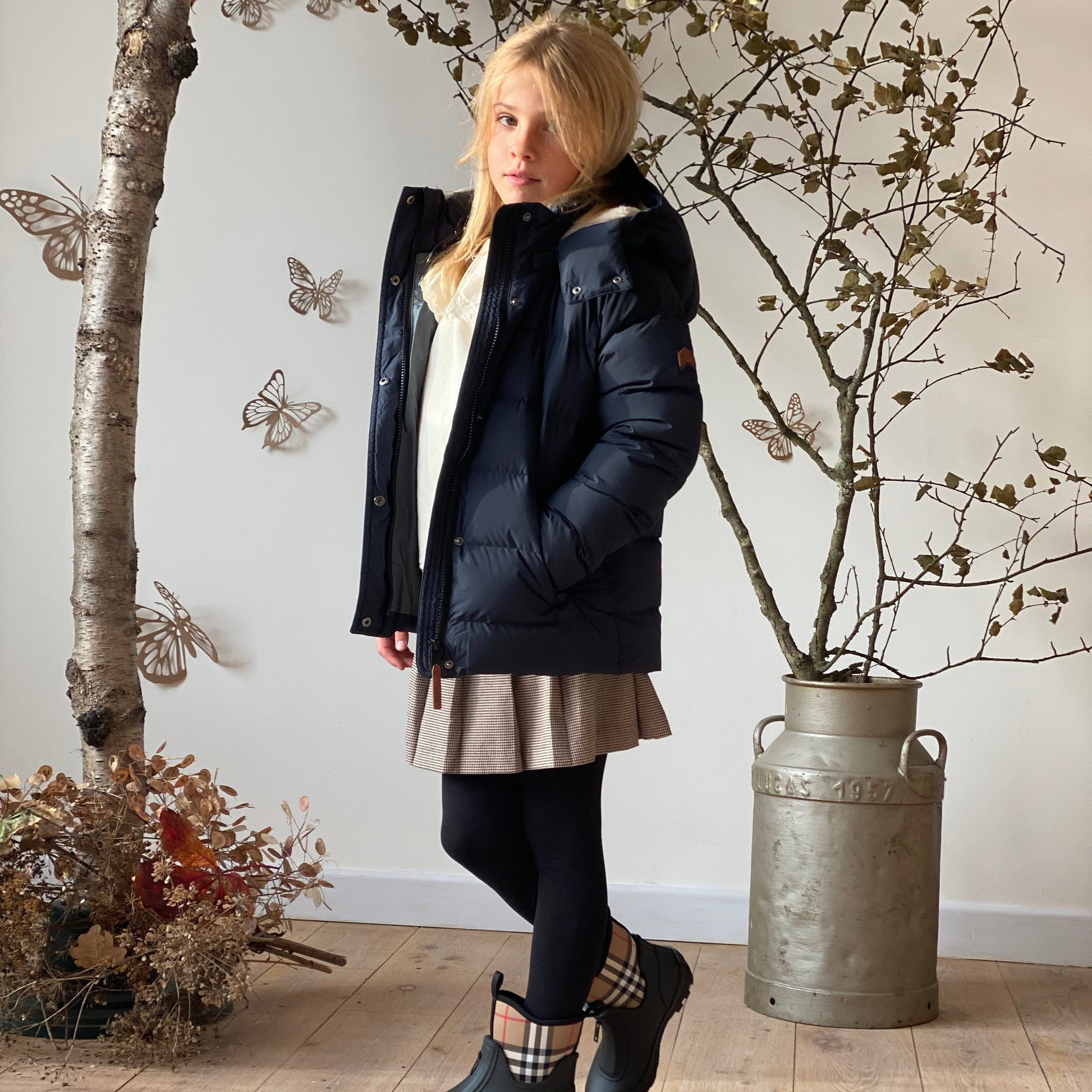 A girl models a dark navy puffer jacket in a styled indoor setting with a wooden floor, a rustic metal canister planter, and butterfly wall decorations.