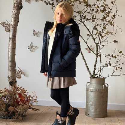 A girl models a dark navy puffer jacket in a styled indoor setting with a wooden floor, a rustic metal canister planter, and butterfly wall decorations.