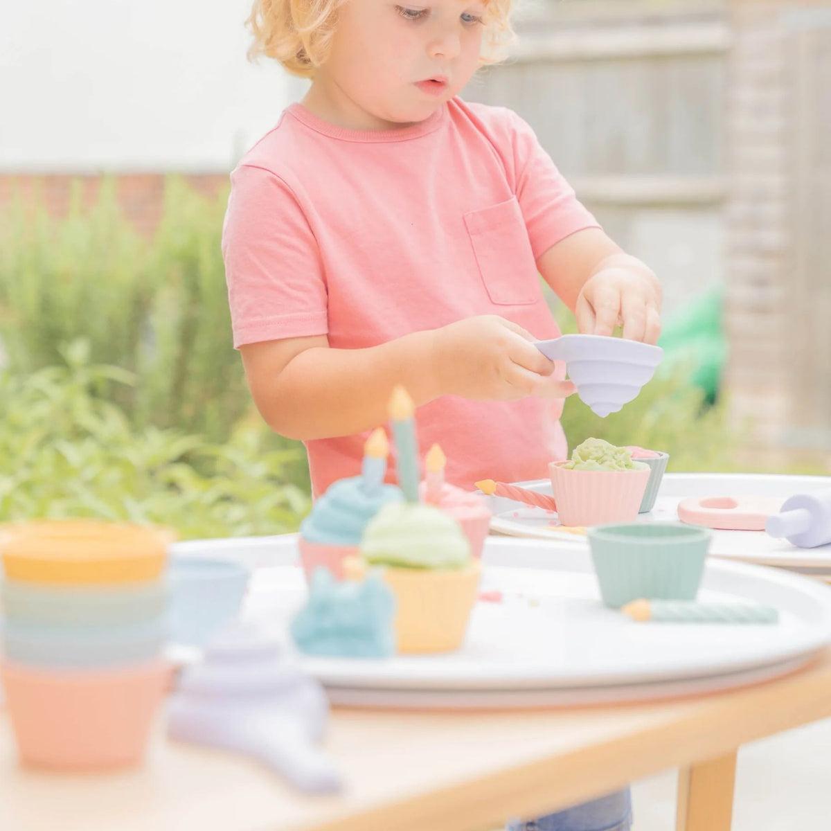 A young child wearing a pink t-shirt playing with the inspire my play cupcake set in pastel-colored tones set on a wooden table outdoors.
