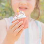 A close-up of a child holding a silicone cupcake liner with pretend icing for sensory play, colorful sprinkles, and a small yellow candle in a pink cupcake liner, with a soft-focus outdoor background.