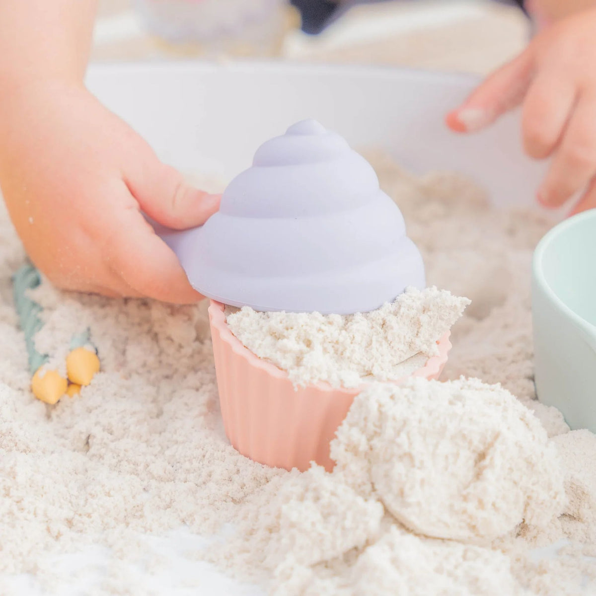 A child's hands pressing a pastel lavender cupcake-shaped mold into light beige kinetic sand, with a pink cupcake liner filled with sand underneath.