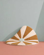 A quilted crescent-shaped floor cushion with alternating tan and cream sunburst stripes, featuring a small pale blue center, set against a two-tone pink and light blue background.