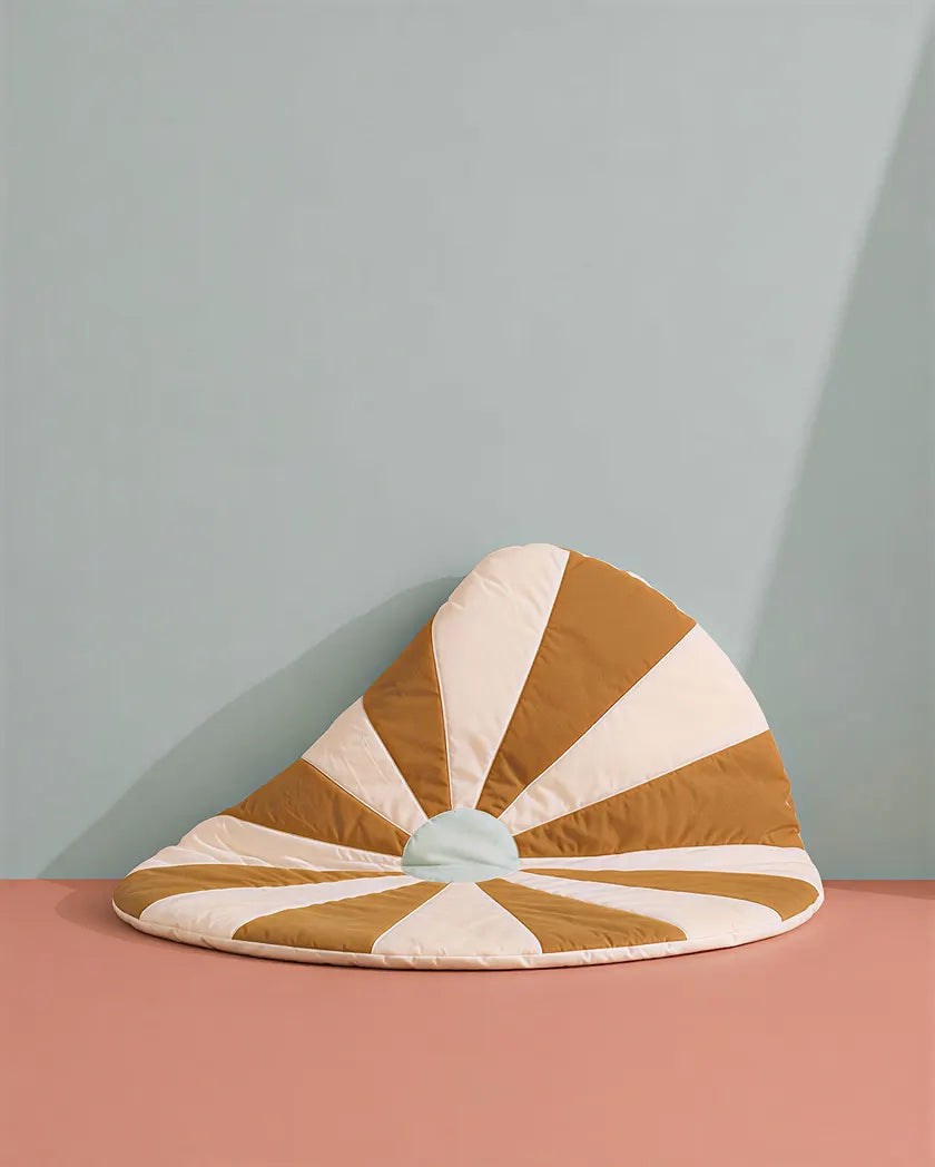 A quilted crescent-shaped floor cushion with alternating tan and cream sunburst stripes, featuring a small pale blue center, set against a two-tone pink and light blue background.