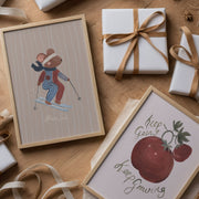Framed artwork with a mouse skiing and an apple, surrounded by wrapped gifts and ribbons on a wooden surface.