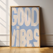 Framed poster with 'GOOD VIBES' text on a light blue background in a room with wooden floor and white walls.