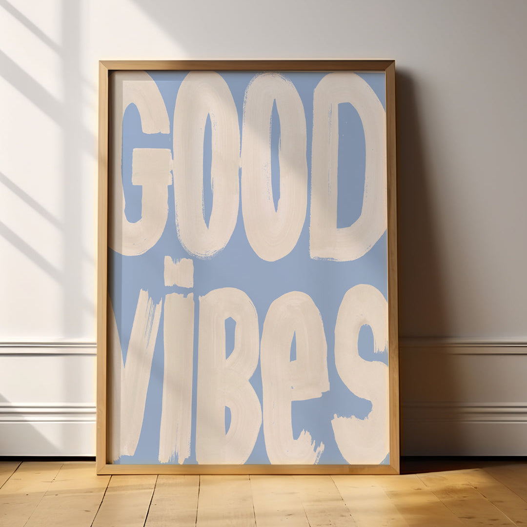 Framed poster with 'GOOD VIBES' text on a light blue background in a room with wooden floor and white walls.