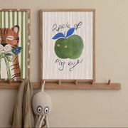Framed artwork with an apple illustration and text on a wall with a decorative hook and plush toy.
