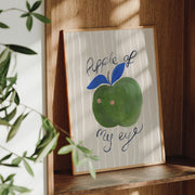 Framed artwork of a green apple with a face and the text 'apple of my eye' on a wooden shelf.