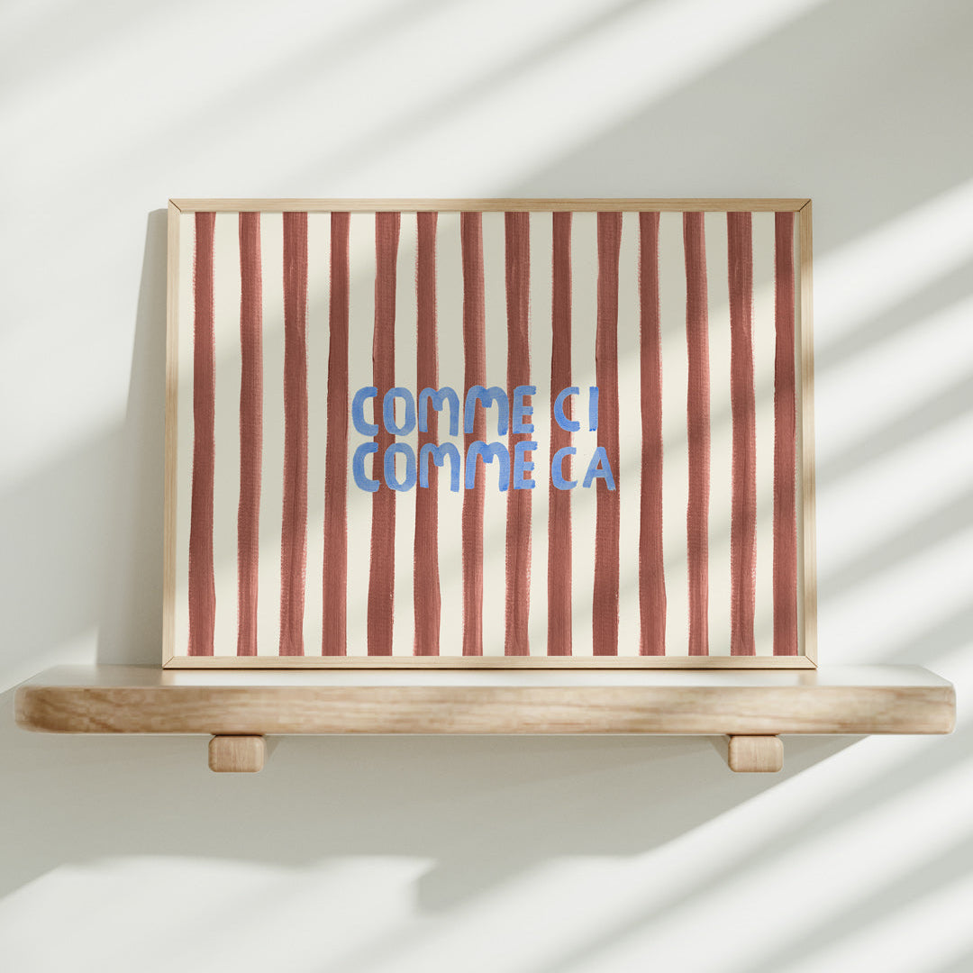 Striped artwork with 'Comme ci, comme ca' text on a wooden shelf against a white wall.