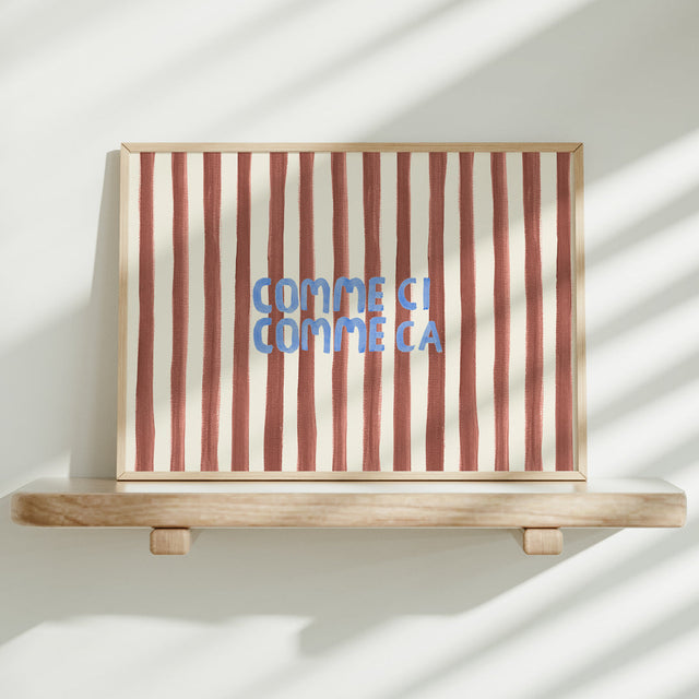 Striped artwork with 'Comme ci, comme ca' text on a wooden shelf against a white wall.