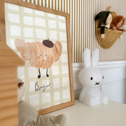 Framed artwork with a croissant character and to word 'Bonjour' on a checkered background, placed on a surface with toys around.