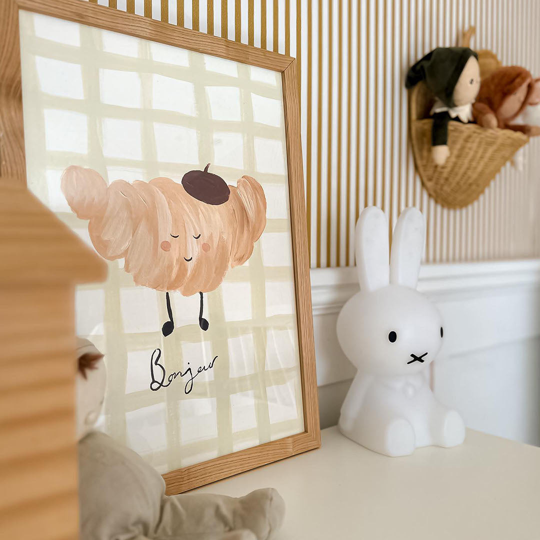 Framed artwork with a croissant character and to word 'Bonjour' on a checkered background, placed on a surface with toys around.