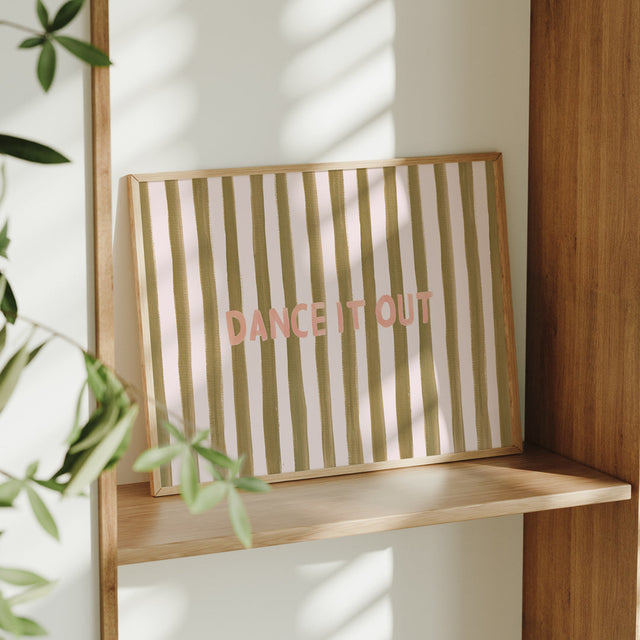 Striped art print with 'DANCE IT OUT' text on a wooden shelf with plant and wall reflection.