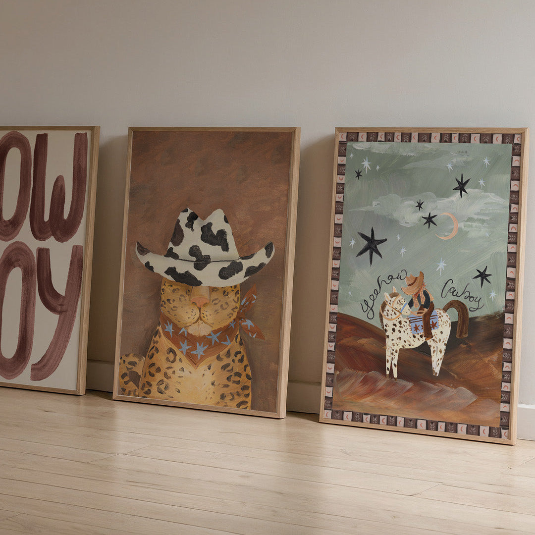 Three framed western themed cowboy artworks on a wooden floor against a plain wall.