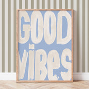 Framed artwork with 'GOOD VIBES' text on a blue background, leaning against a striped wall.