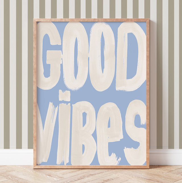 Framed artwork with 'GOOD VIBES' text on a blue background, leaning against a striped wall.