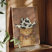 Framed artwork of a leopard wearing a cowgirl hat on a wooden shelf.