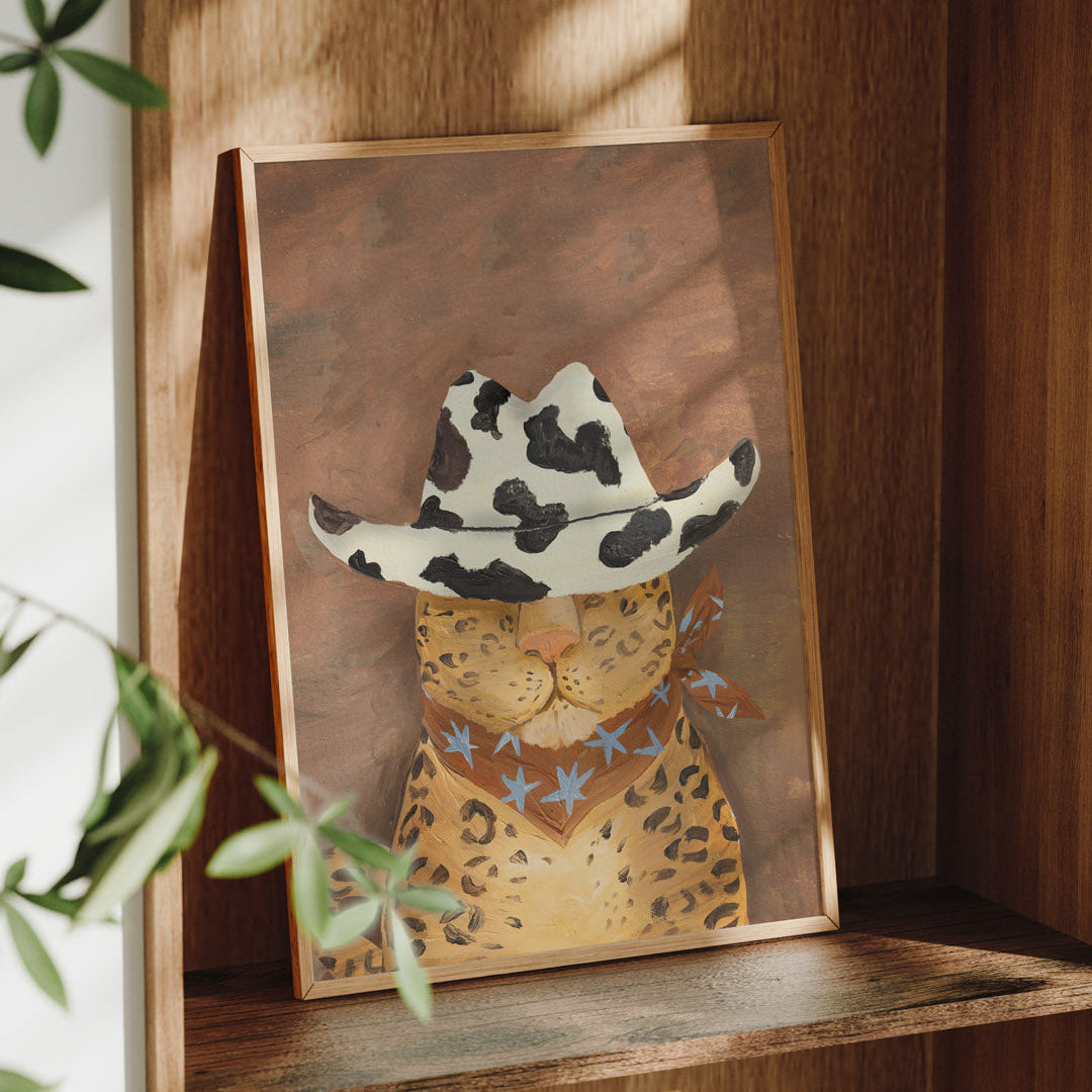 Framed artwork of a leopard wearing a cowgirl hat on a wooden shelf.