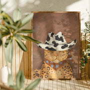 Framed artwork of a leopard wearing a cowboy hat with star patterns, placed on a surface with plants in the background.