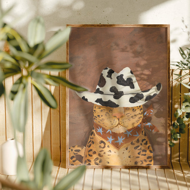 Framed artwork of a leopard wearing a cowboy hat with star patterns, placed on a surface with plants in the background.