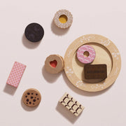 A set of wooden toy food pieces arranged on a pale pink background, including a round tray with a pink donut and a brown Bourbon chocolate piece, plus separate cookies, a pink patterned rectangle, and striped candy pieces.