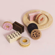 A set of wooden toy sandwich cookies and pastries arranged on a pale pink background, including a donut, chocolate bar, layered cookie, Oreo-style cookie, and a round tray with pink striped cards.