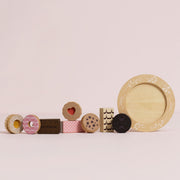 A set of wooden toy food and tableware pieces arranged on a pale pink background, including donut-shaped pieces, a chocolate bar labeled DUBROUN, cookies, a plate, and a bowl.