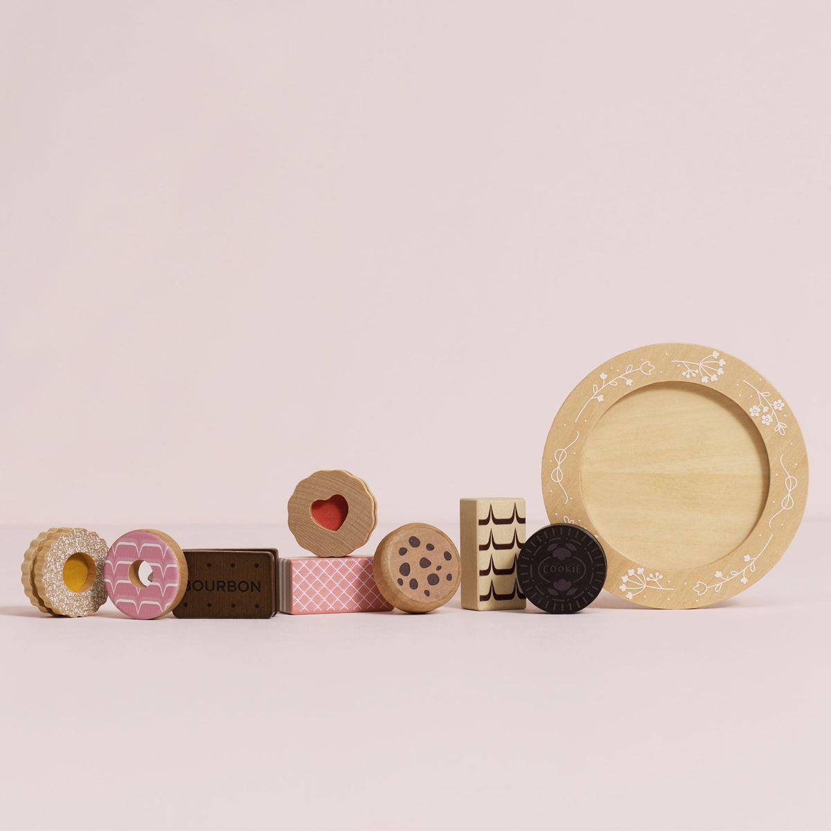 A set of wooden toy food and tableware pieces arranged on a pale pink background, including donut-shaped pieces, a chocolate bar labeled DUBROUN, cookies, a plate, and a bowl.