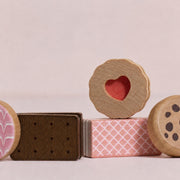 A set of wooden toy sandwich cookies and cake blocks, including a heart-shaped biscuit on top of a pink patterned cake base, with other toy cookies partially visible on both sides, set against a solid pale pink background.