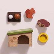 A set of handcrafted wooden toys arranged on a light beige background, including a small house with a green roof, a squirrel figure, a hedgehog figure, a round yellow bowl, and a dark brown piece with two round knobs.