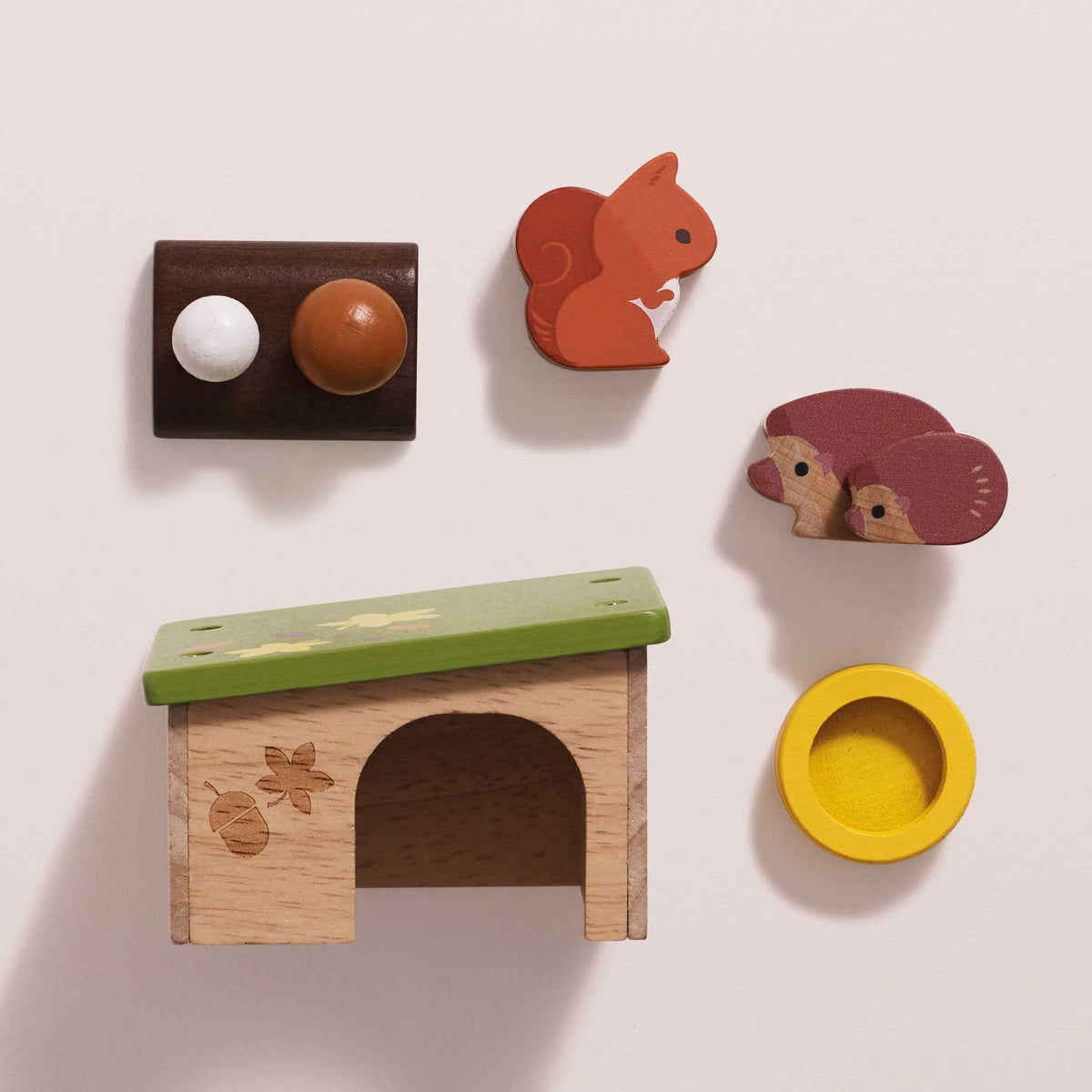 A set of handcrafted wooden toys arranged on a light beige background, including a small house with a green roof, a squirrel figure, a hedgehog figure, a round yellow bowl, and a dark brown piece with two round knobs.