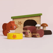 A set of wooden children's toys on a pale pink background, including a small house with a green roof, a squirrel figure, a hedgehog figure, two mushrooms, and a yellow bowl with a flower.