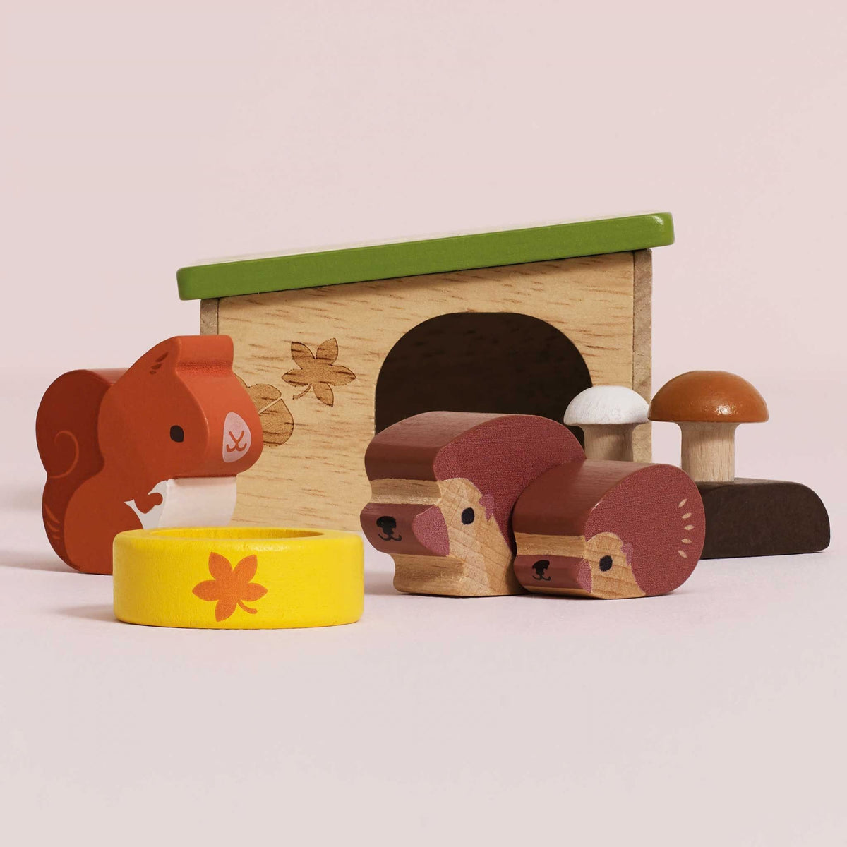 A set of wooden children's toys on a pale pink background, including a small house with a green roof, a squirrel figure, a hedgehog figure, two mushrooms, and a yellow bowl with a flower.