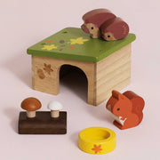 A wooden toy play set featuring a small house with a green roof, two squirrel figures on top and beside it, two mushrooms on a dark base, and a yellow ring with a flower, all photographed on a soft light pink background.