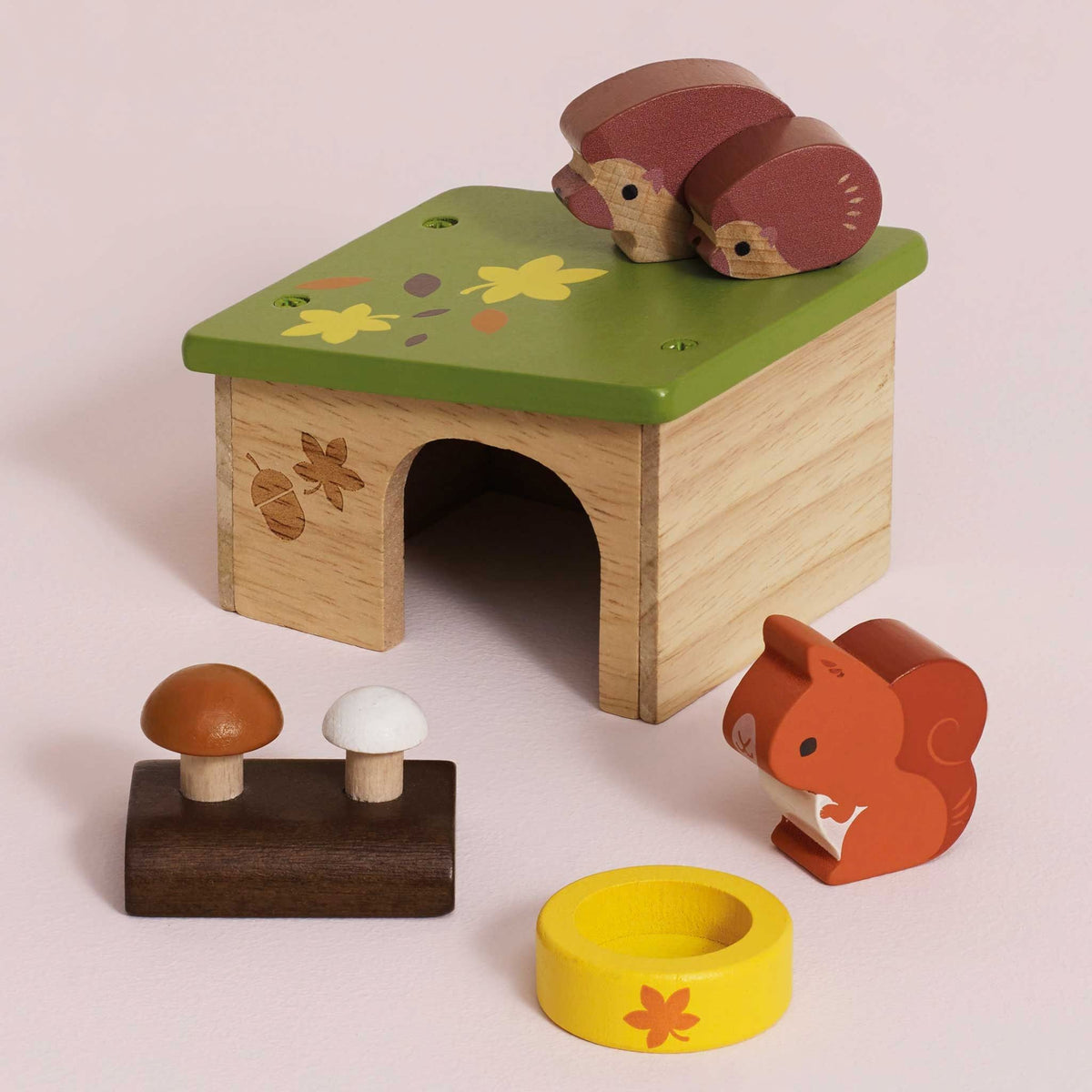 A wooden toy play set featuring a small house with a green roof, two squirrel figures on top and beside it, two mushrooms on a dark base, and a yellow ring with a flower, all photographed on a soft light pink background.