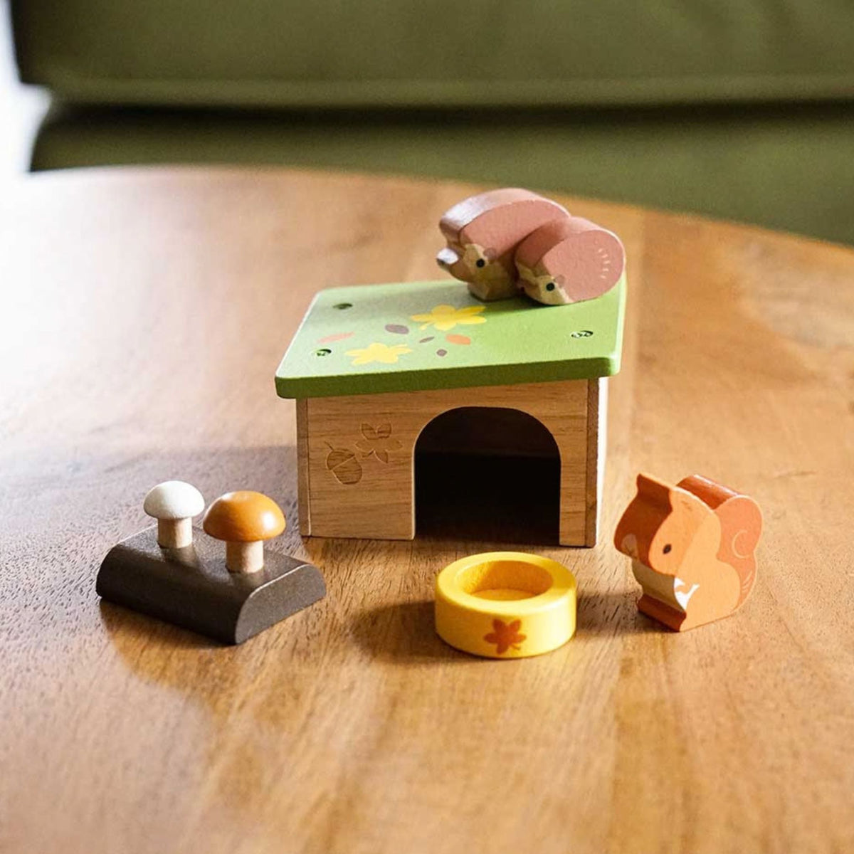 A wooden toy set featuring a small house with a green roof, two squirrels, two mushrooms, and a yellow ring, displayed on a wooden table.