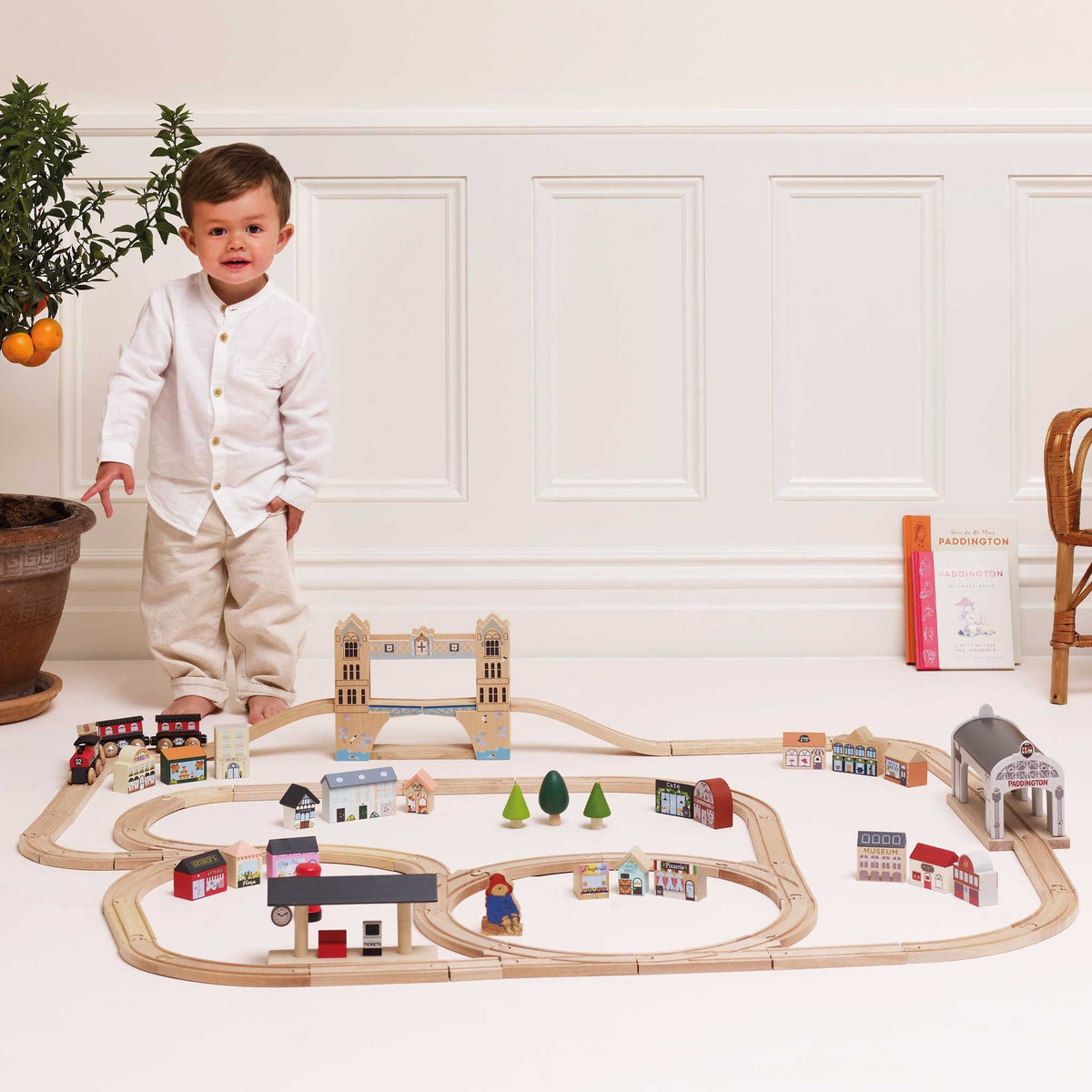 A toddler stands beside a large wooden train track set with houses, bridges, signals, and a train in a bright indoor room with white paneled walls.