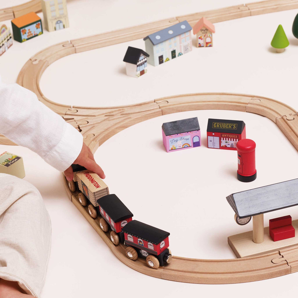 A wooden toy train set with curved tracks on a light beige floor, featuring a red and black locomotive pulling small cargo cars. A child's hand in a white sleeve is guiding the train, with miniature houses, shops, and a station platform nearby.