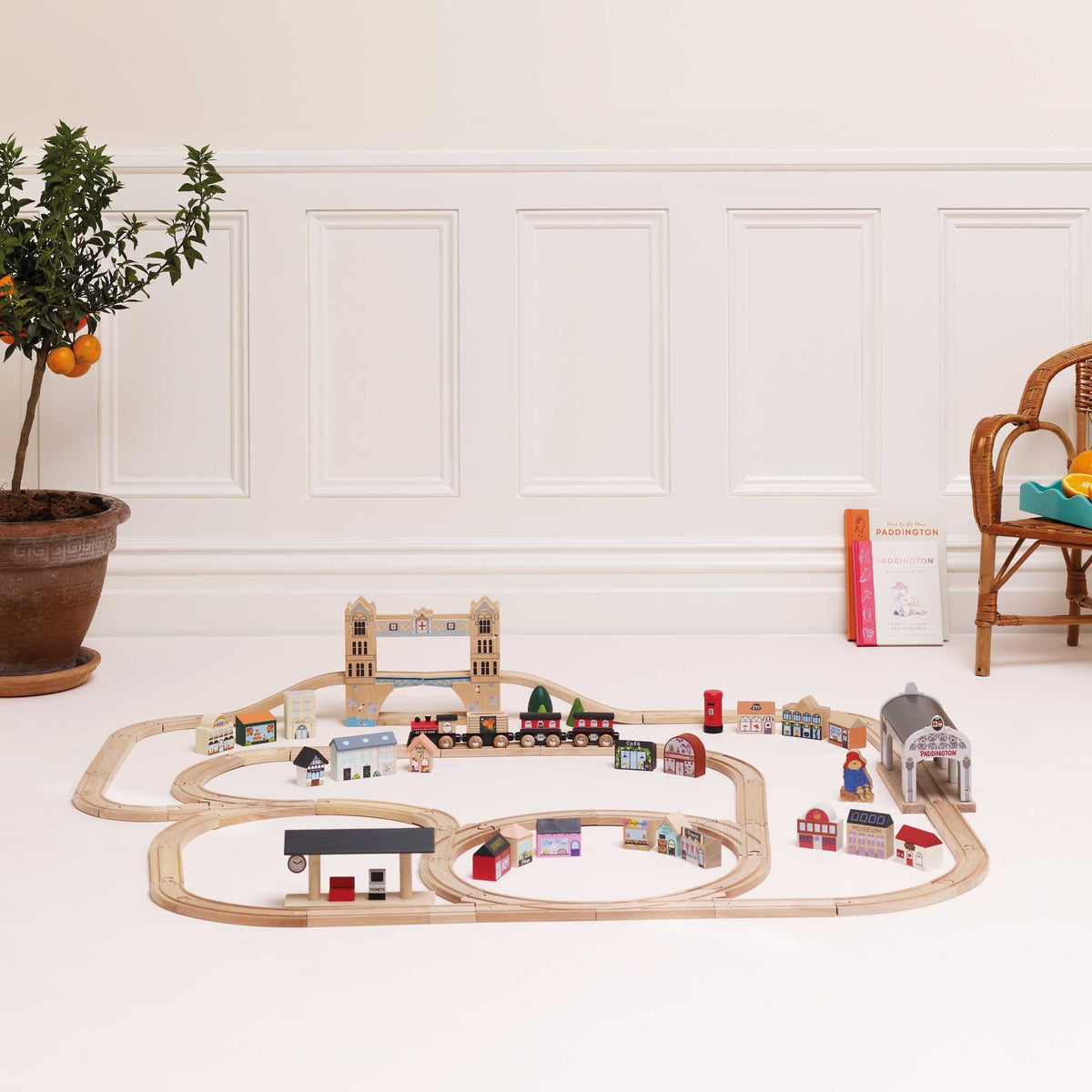 A wooden toy train set arranged on a white floor in a bright room, with a potted plant on the left and a wicker chair on the right. The set includes light wood tracks, a train, bridges, tunnels, and small colorful house and landscape pieces.