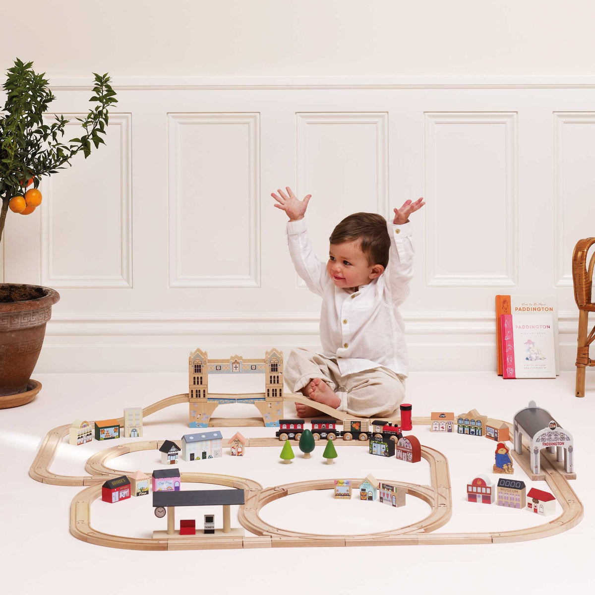 A toddler sits cross-legged on a white floor, surrounded by a wooden toy train set with tracks, small houses, and a bridge. The child is wearing a white long-sleeve top and beige pants, with a white paneled wall and a potted plant in the background.