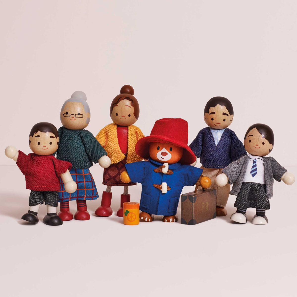 A set of six handcrafted wooden toy figurines representing a family, including a father, mother, three children, and two grandparents, with Paddington Bear in the center holding a suitcase.