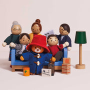 A set of wooden Paddington Bear family figurines and accessories, including a blue sofa, green lamp, and small blocks, photographed on a clean off-white background.