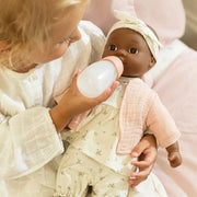 A close-up lifestyle shot of a young child in a white outfit feeding a baby doll with a white bottle. The doll is dressed in a cream floral outfit with a pink muslin vest and a cream bow headband, resting on a soft pink bed.