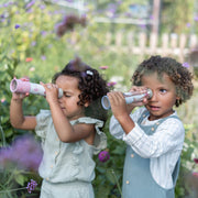 Two young children outdoors in a flower garden, each holding a pink and white paper telescope and looking through it.