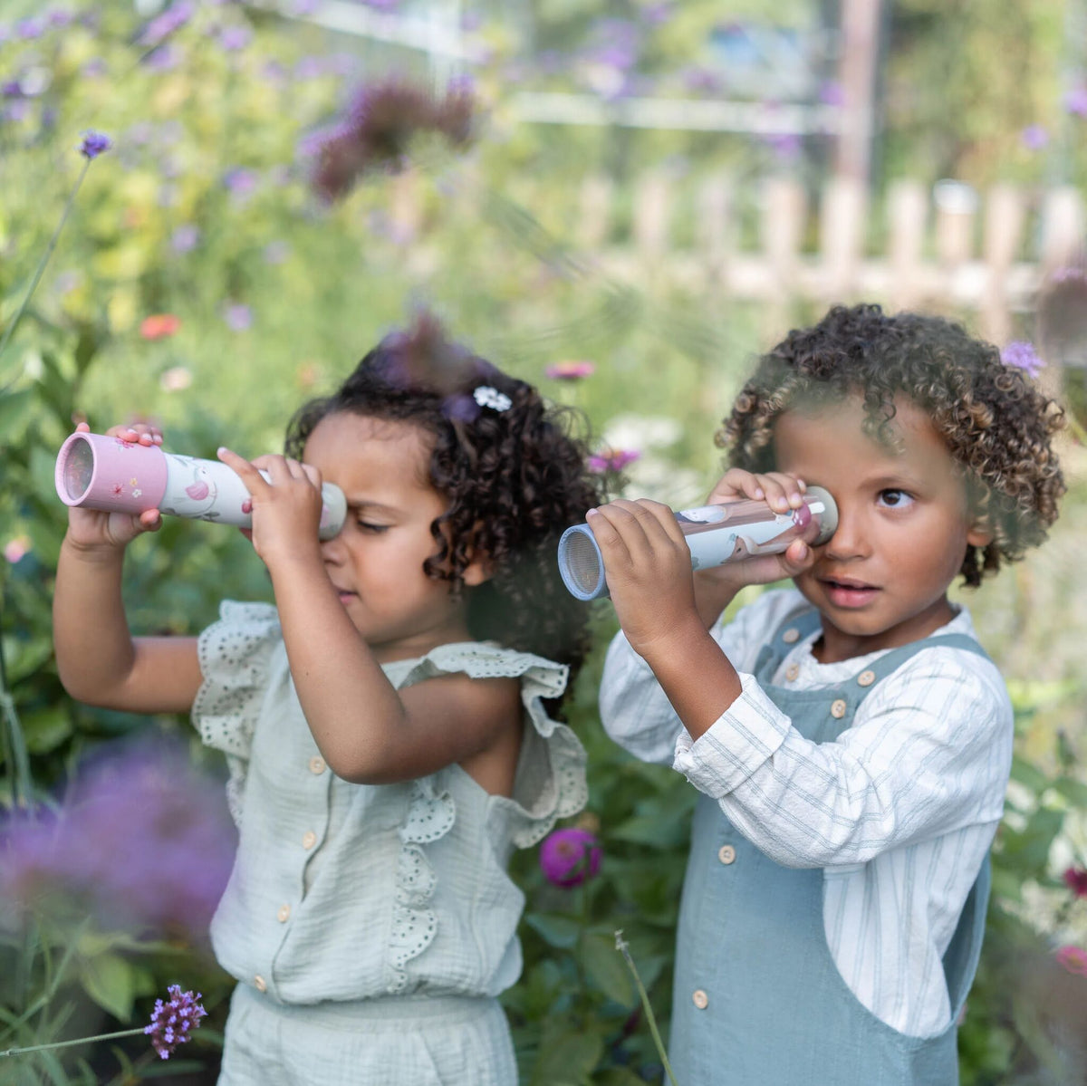Two young children outdoors in a flower garden, each holding a pink and white paper telescope and looking through it.