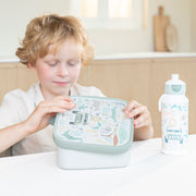 A young child with blonde hair sits at a table, holding a Little Dutch light green and white patterned lunch box. A matching water bottle stands to the right on the white tabletop.