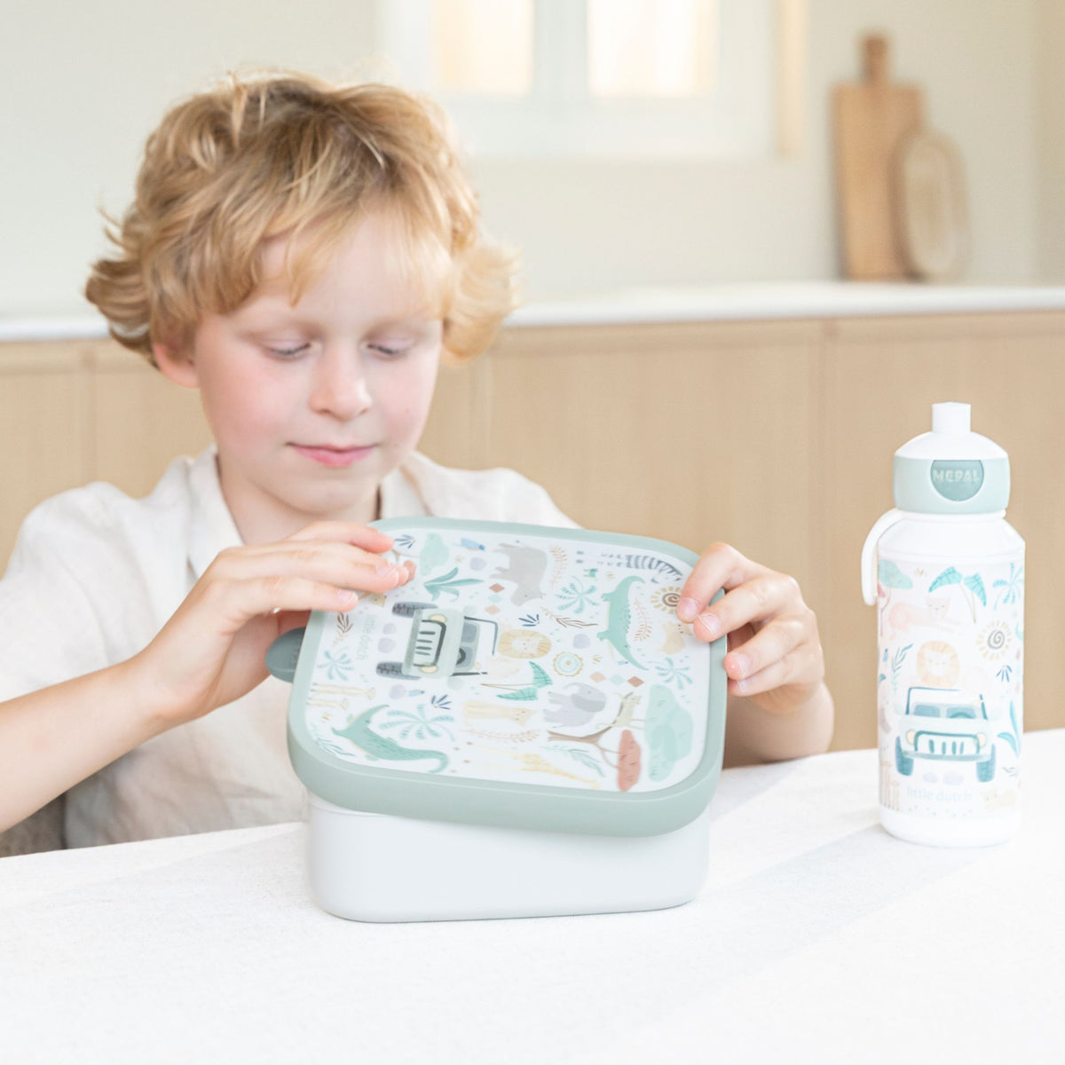 A young child with blonde hair sits at a table, holding a Little Dutch light green and white patterned lunch box. A matching water bottle stands to the right on the white tabletop.
