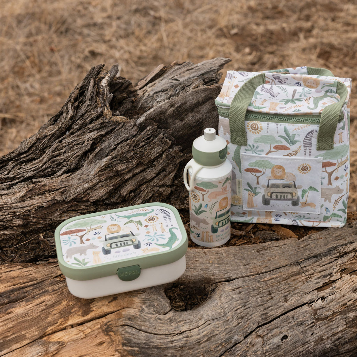 A coordinated Little Dutch picnic-style set on weathered logs outdoors, including a rectangular lunch box, a reusable bottle, and a soft-sided tote bag. The items share a safari-themed print with green handles and trim, plus illustrated animals and vehicles on a light background.